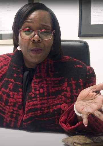 A woman in a red and black patterned coat gestures while sitting at a desk.