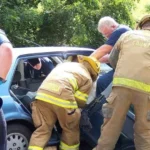Firefighters and rescue personnel assist a person inside a blue sedan after an apparent accident.
