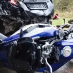 Close-up of a motorcycle accident scene showing a blue motorcycle lying on its side in front of a heavily damaged black car