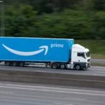 A white semi-truck with a bright blue Amazon Prime trailer drives on a highway, passing greenery in the background.