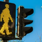 Pedestrian crossing signal lit yellow beside a traffic light against a clear blue sky.