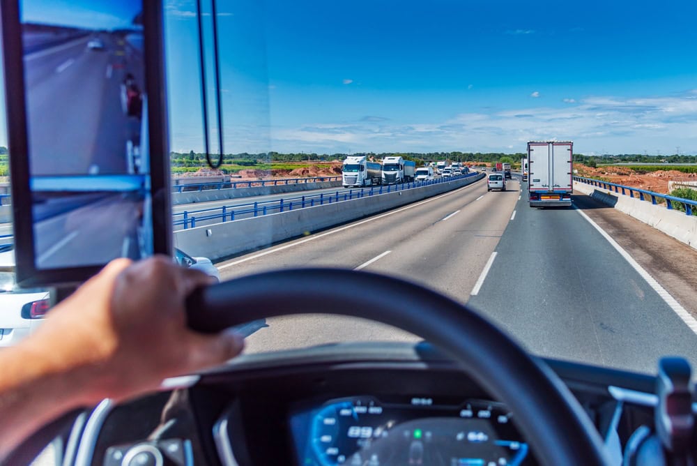 View from truck driver's seat showing hands on steering wheel while driving on busy Texas highway with multiple commercial trucks, illustrating hours of service regulations and road safety